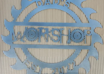A blue metallic sign with the words _DAD'S WORKSHOP EST. 2012_ surrounded by saw and gear shapes.